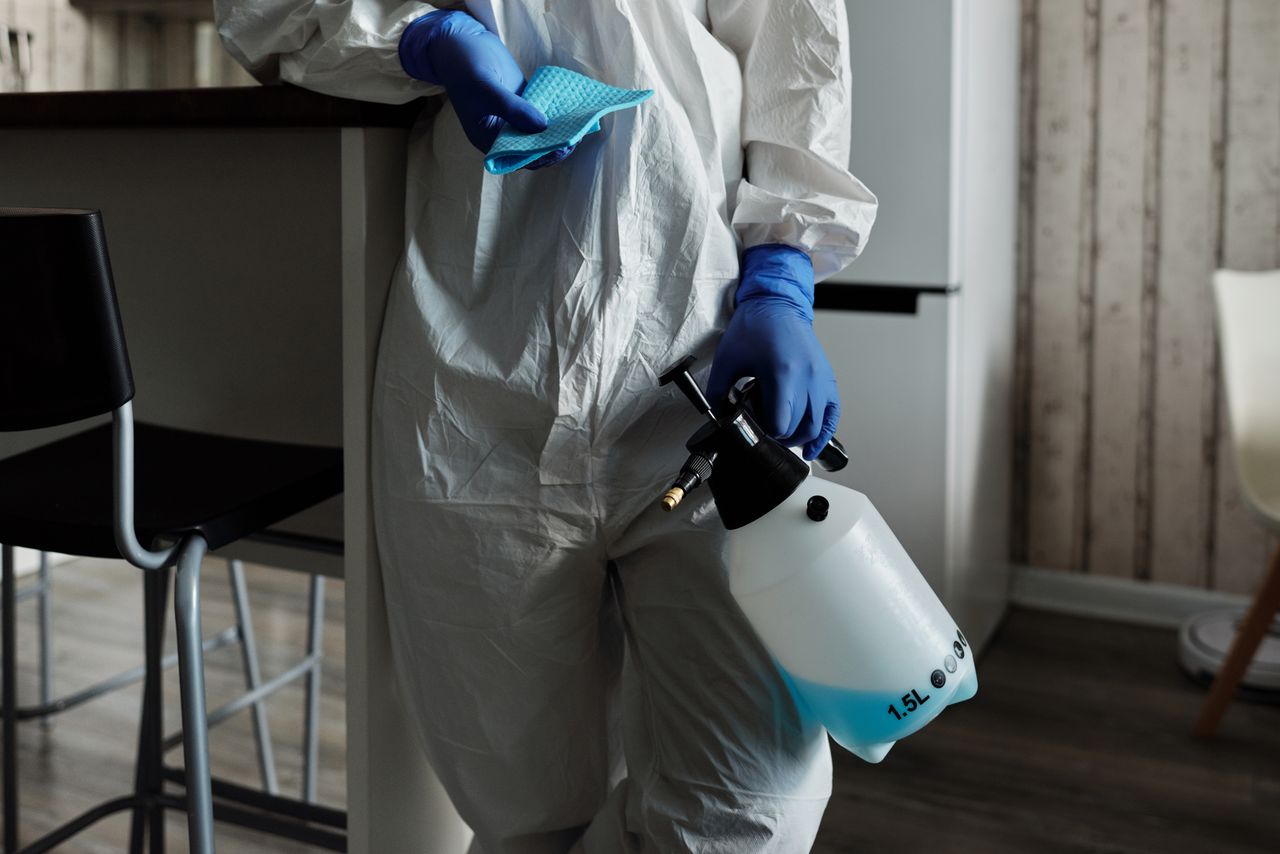 Individual in PPE holding a disinfectant spray bottle indoors for cleaning.