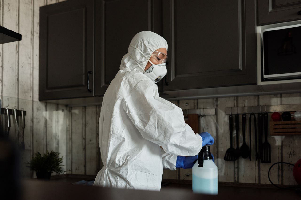 Healthcare worker in PPE uses disinfectant spray in a kitchen setting during COVID-19 pandemic.